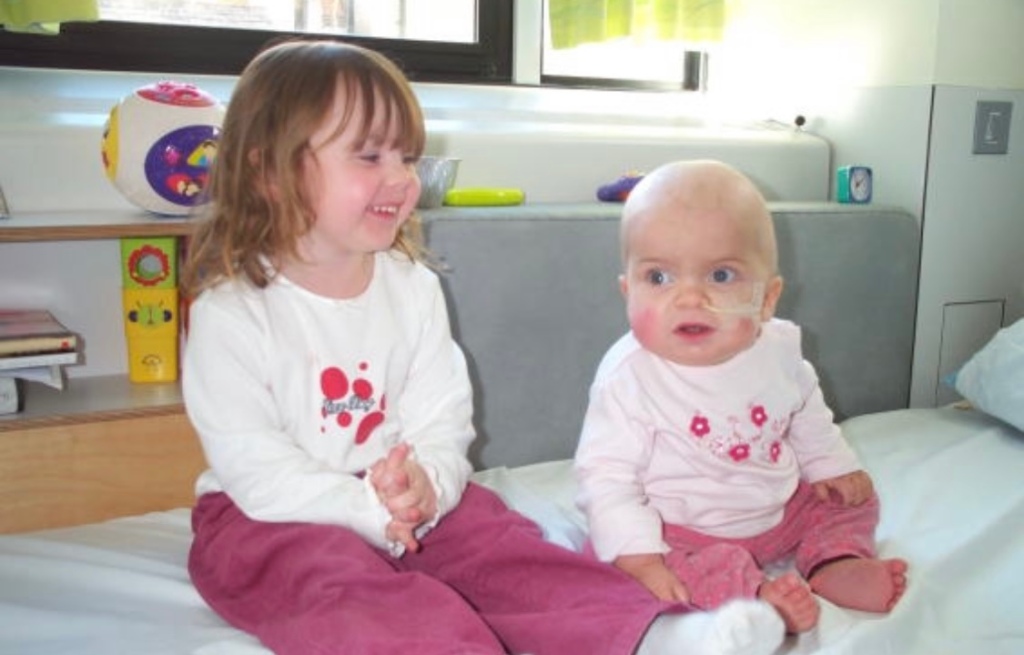 Sophie and Amy sitting on a hospital bed, both wearing pink trousers and a white top with flowers on, Amy is 3 and Sophie is 2. Amy has short brown hair and Sophie does not have hair. Sophie has an NG tube in. Amy is smiling and Sophie is looking into the distance.